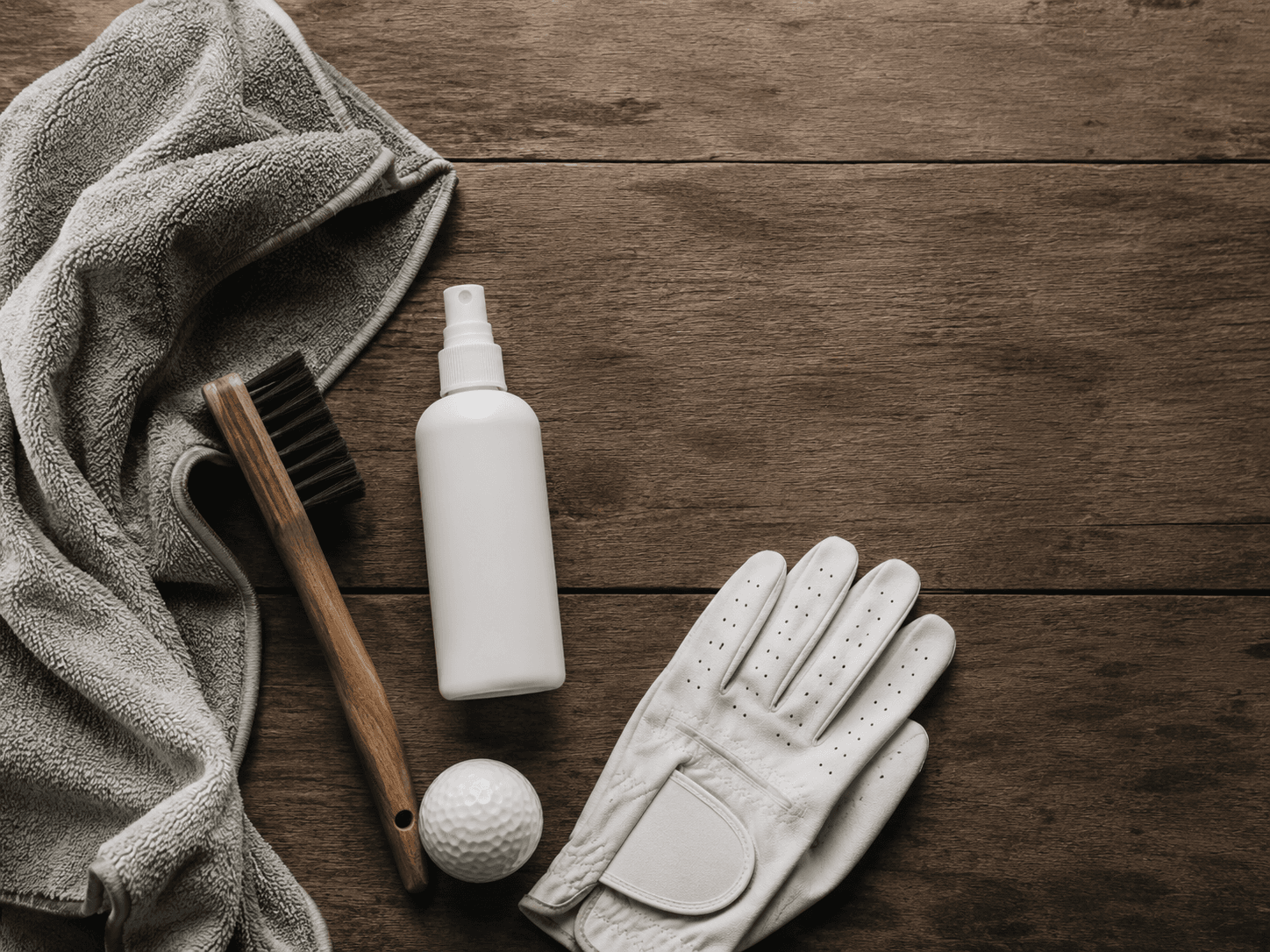 Golf cleaning gear flat lay: towel, brush, spray bottle, glove, and golf ball on a wood surface